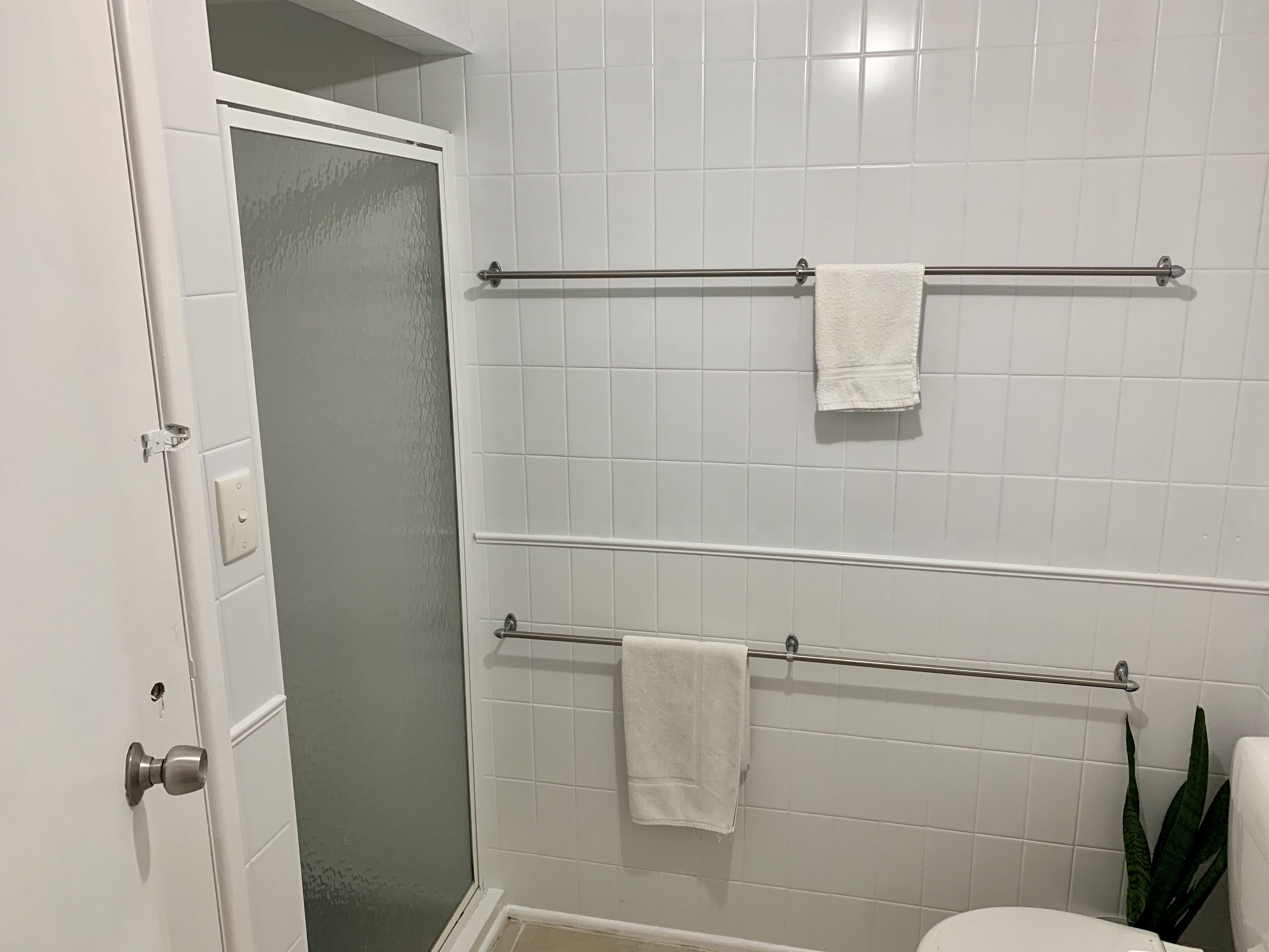 A bathroom with white tiled walls, a glass shower door, and two towel bars with white towels.
