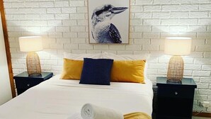 A neatly made bed with a white duvet, two pillows in yellow and blue, and a rolled towel on the bed. There are two bedside tables with lamps and a framed picture on the wall.