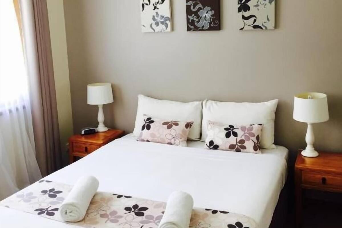 A neatly made bed with floral bedding and two pillows, flanked by bedside tables with lamps, in a room with a ceiling fan and two wall-mounted artworks.