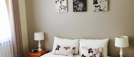 A neatly made bed with floral bedding and two pillows, flanked by bedside tables with lamps, in a room with a ceiling fan and two wall-mounted artworks.