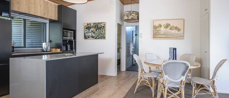 A modern kitchen with a central island, dining area with wicker chairs, and artwork on the walls.