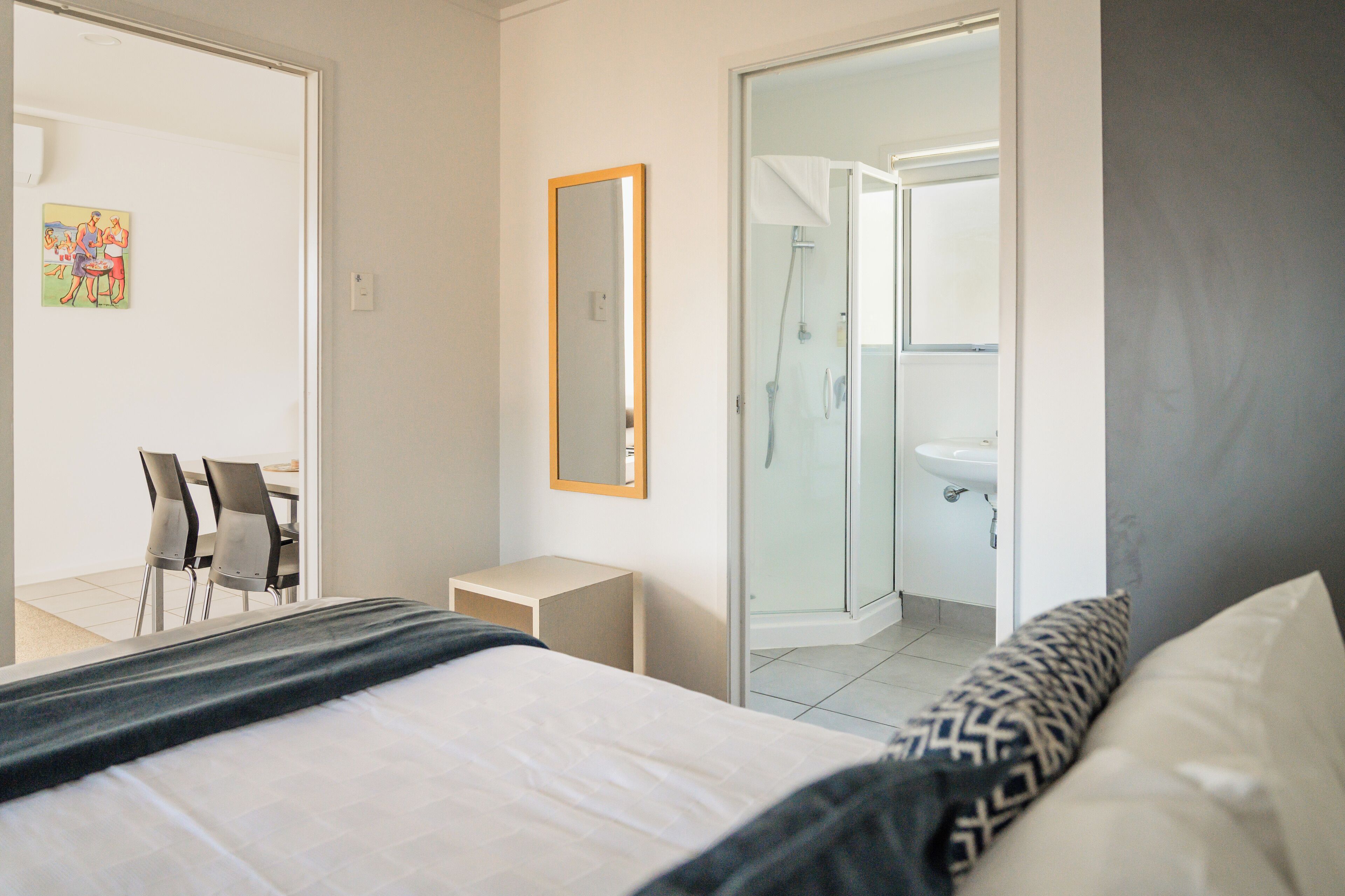 A hotel room with a bed, a bedside table, a chair, a mirror, and a bathroom with a shower and sink.