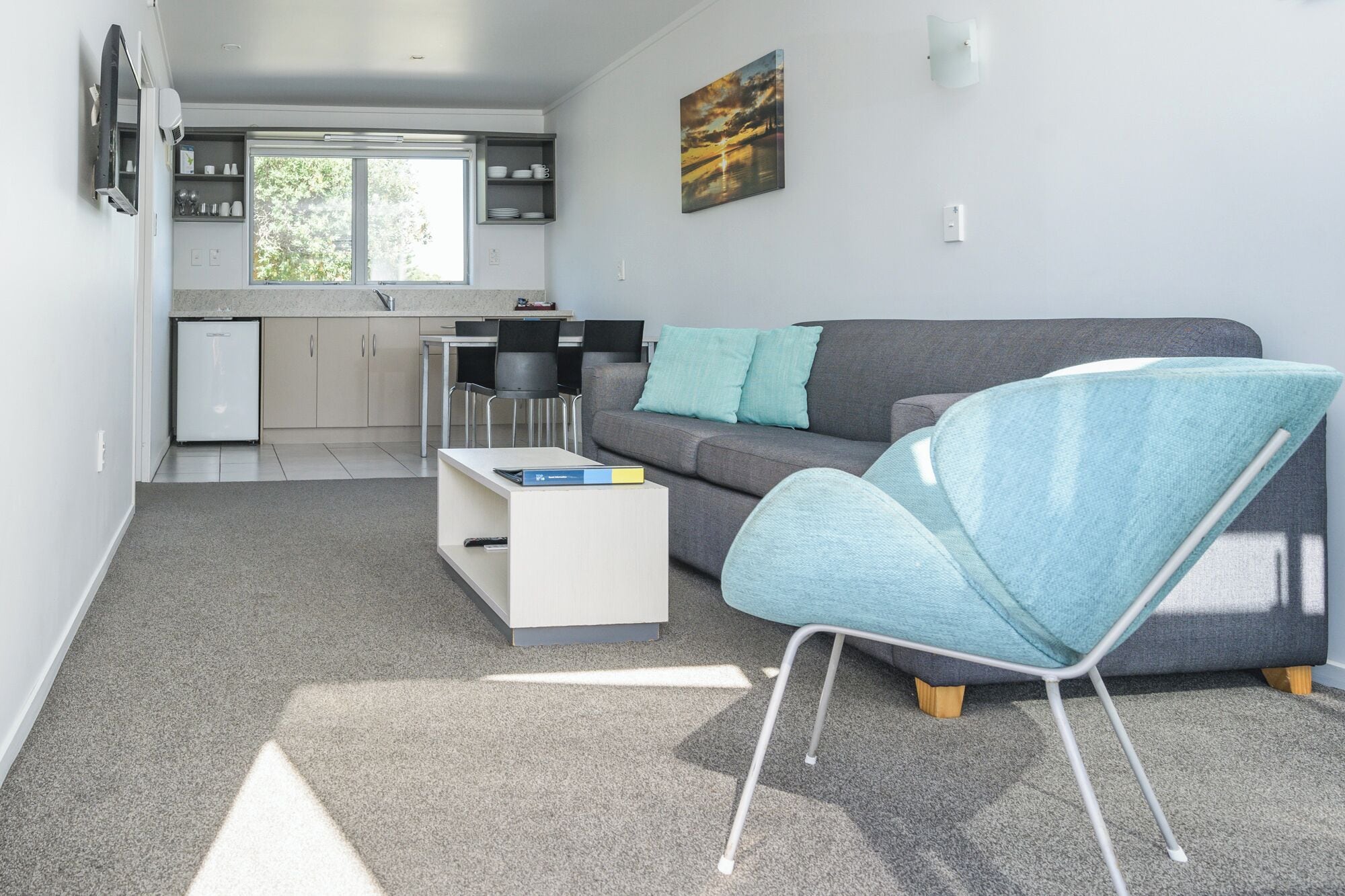 Una sala moderna con un sofá gris, un sillón azul y una pequeña mesa de centro blanca. Se ve una cocina con ventana y lavamanos al fondo.