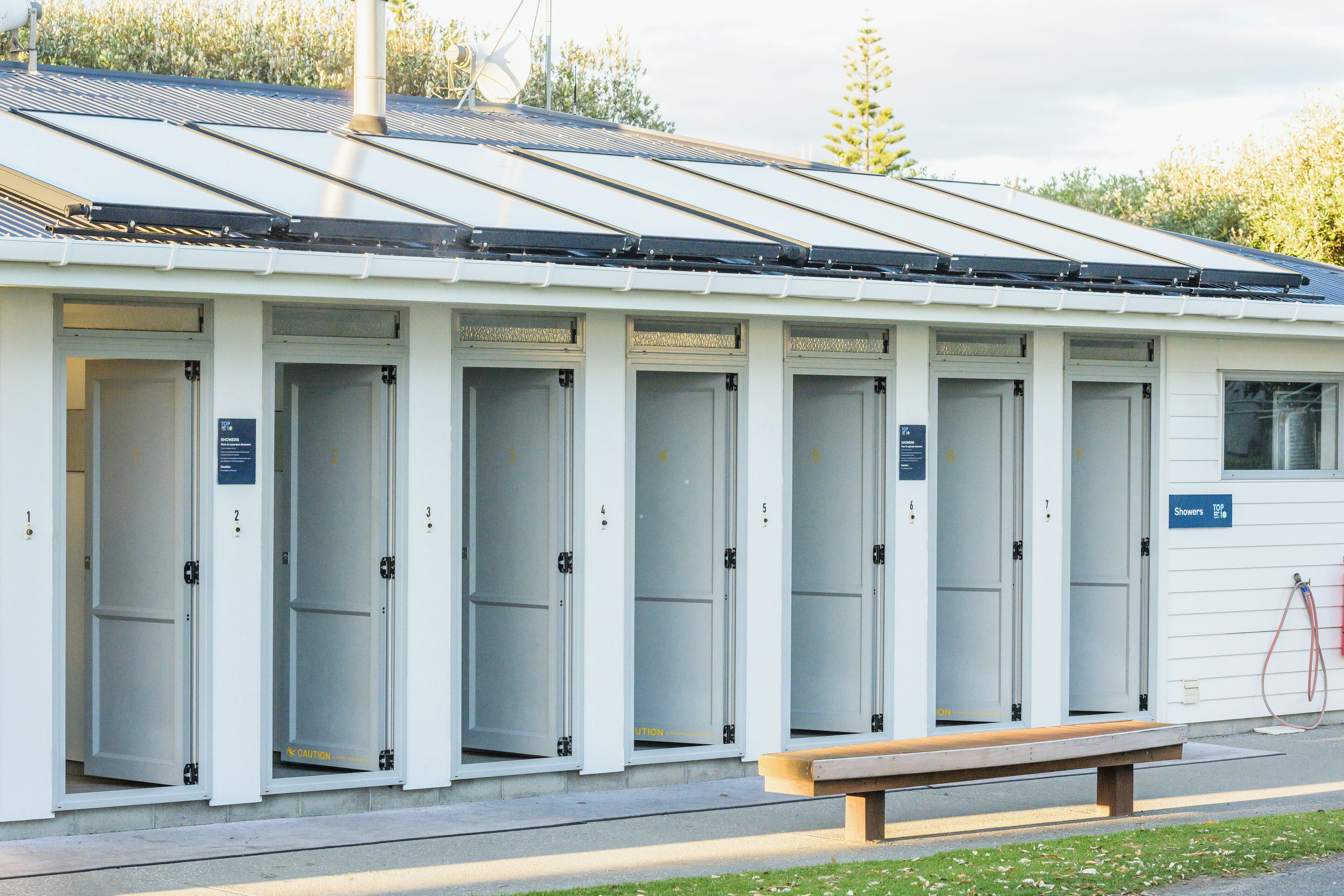 Une rangée de toilettes publiques mobiles blanches, dotées de portes numérotées, avec un banc à l’extérieur et un panneau solaire sur le toit.