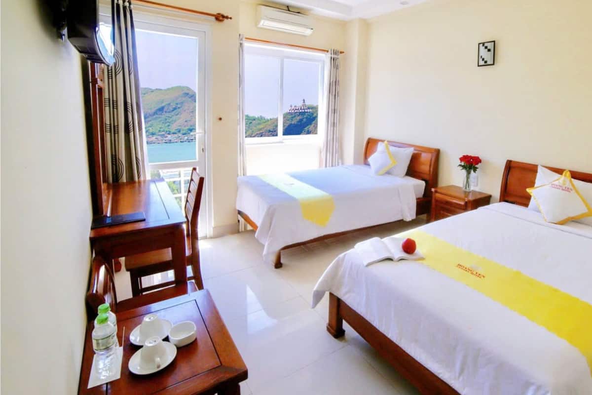 Une chambre d’hôtel avec deux lits, un bureau en bois avec une chaise, une petite table avec une bouteille et des verres, et une vue sur un paysage montagneux par la fenêtre.