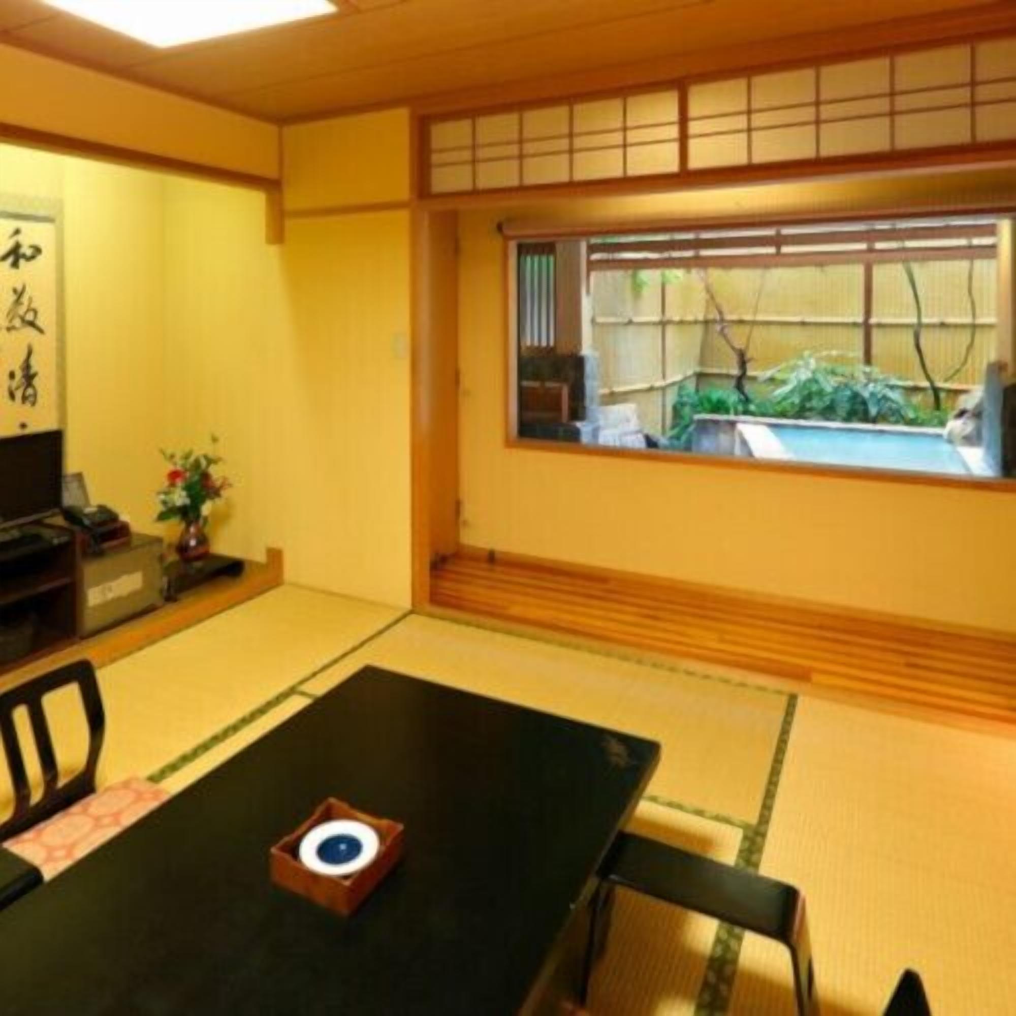 Ein traditioneller japanischer Raum mit Tatami-Böden, einem niedrigen Tisch mit schwarzer Matte, einem Holzstuhl, einem kleinen Tisch mit Dekoration und einem Fenster mit Blick auf Grünflächen.