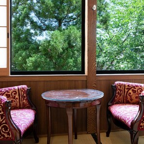 A room with two wooden chairs with cushioned seats and backs, a small round table, and a large window with a view of trees. - Hot Springs Inn 'unzen Iwaki Ryokan' (Unzen)