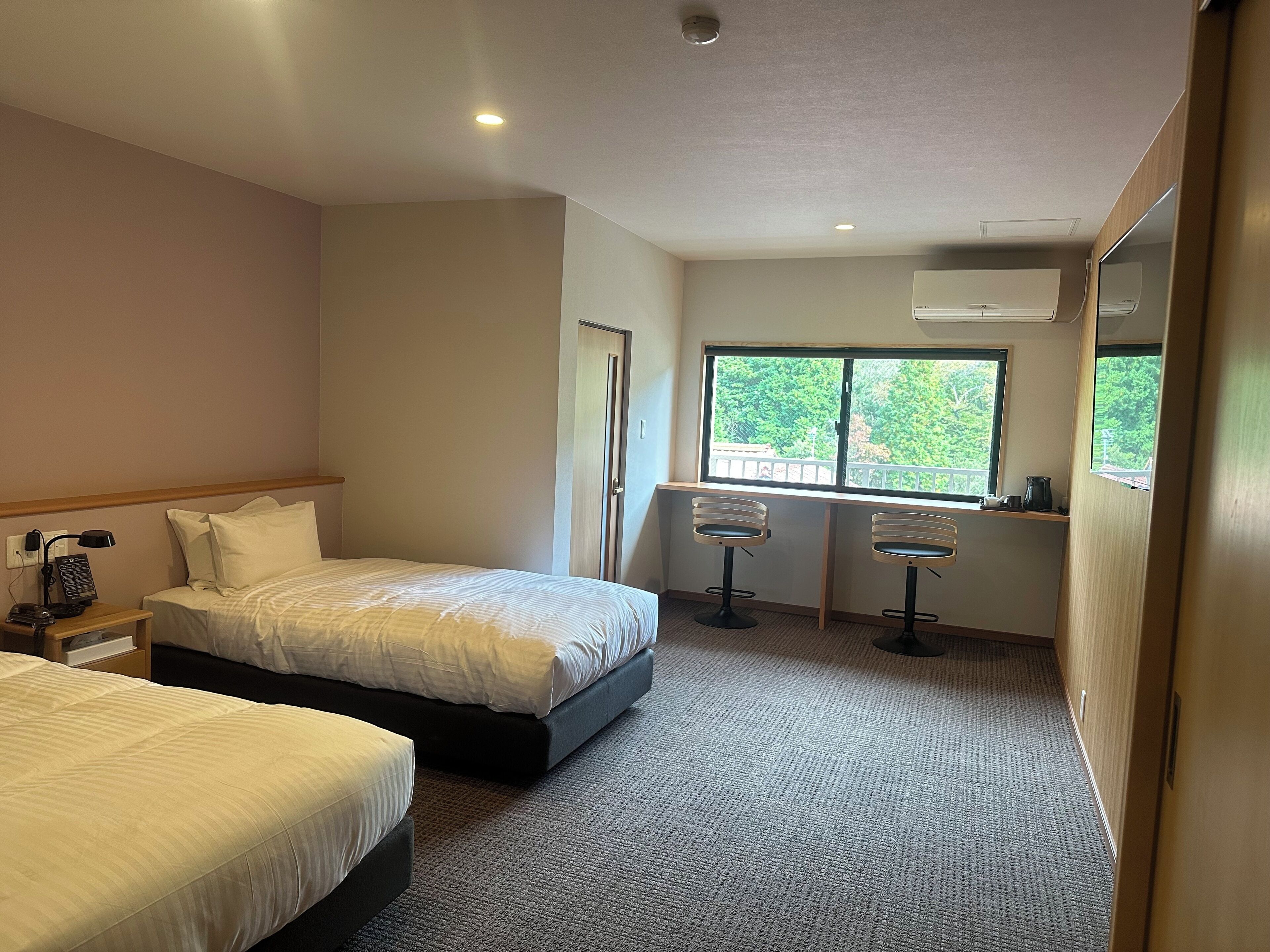 Ein Hotelzimmer mit zwei Betten, einem Schreibtisch mit zwei Barhocker und einem Fenster mit Blick ins Grüne, sowie einer Klimaanlage.