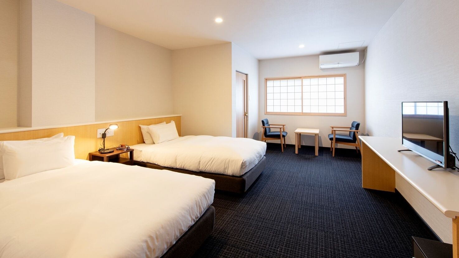Ein Hotelzimmer mit zwei Betten, einem Schreibtisch, einem Stuhl, einem Fernseher und einem Fenster.
