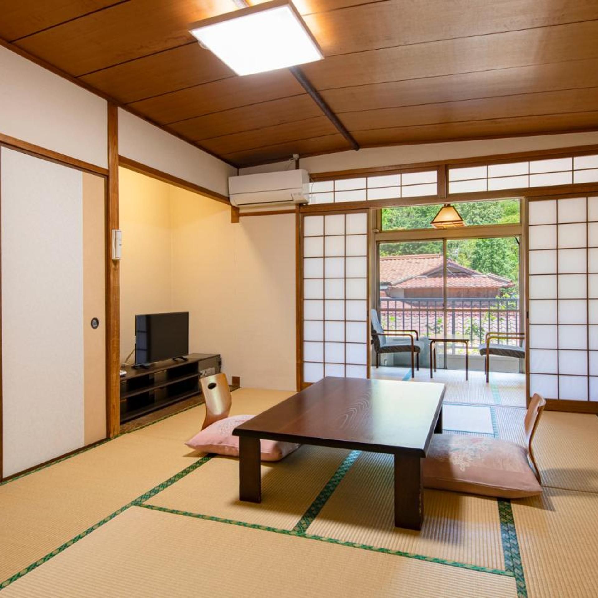 A traditional Japanese room with tatami flooring, a low table, a television, and sliding doors leading to an outdoor area.