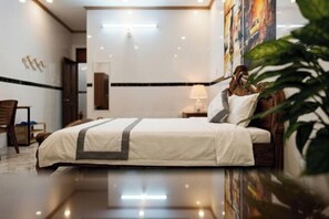 A hotel room with a bed, a nightstand, a lamp, a chair, and a large painting on the wall. - CT Morning Hotel (Can Tho)