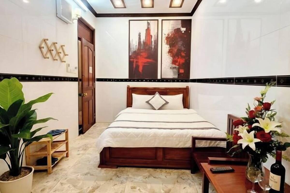 Une chambre d’hôtel avec un lit, une tête de lit en bois, une table de chevet, une plante en pot et des œuvres d’art accrochées au mur.