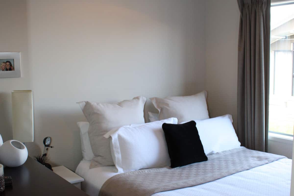 Una cama bien hecha con sábanas blancas y almohada negra, una mesita de noche con lámpara, y una ventana con cortinas.
