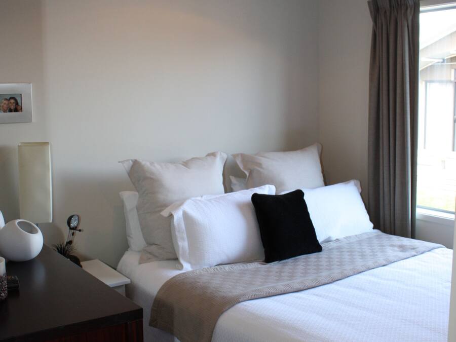 A neatly made bed with white linens and a black pillow, a bedside table with a lamp, and a window with curtains.