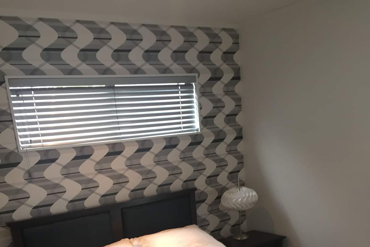 Un dormitorio con cama, almohadas, mesita de noche, lámpara y ventana con persianas.