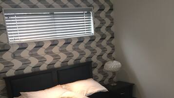A bedroom with a bed, pillows, a nightstand, a lamp, and a window with blinds.