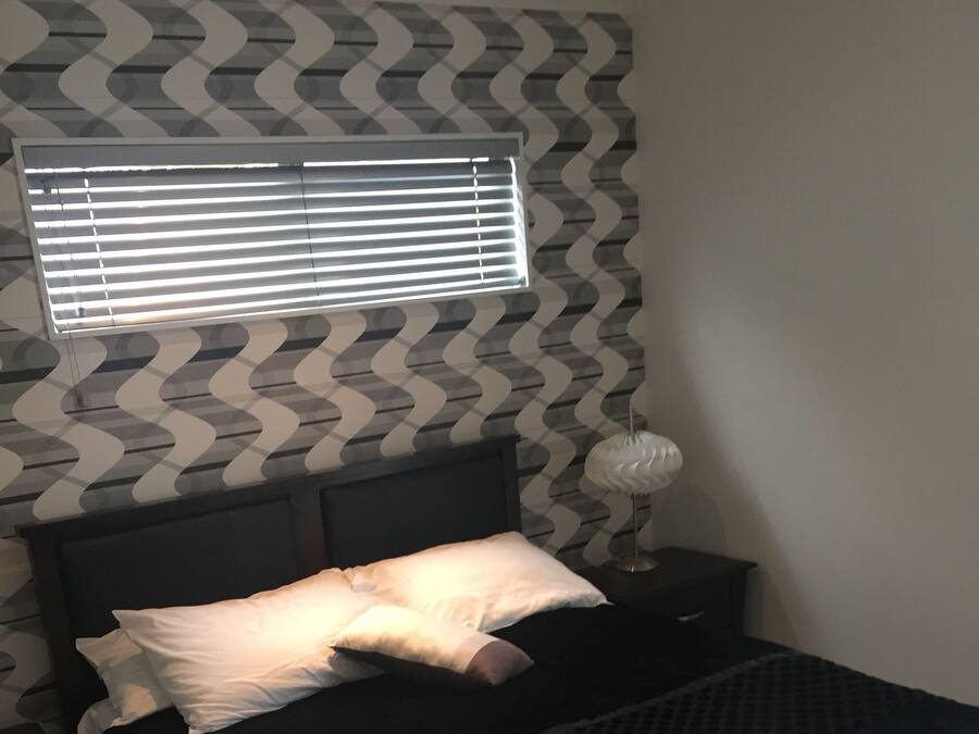 A bedroom with a bed, pillows, a nightstand, a lamp, and a window with blinds.