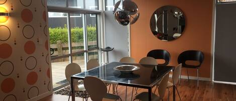 A modern dining area with a glass table, six chairs, a large mirror, and a view of greenery outside.