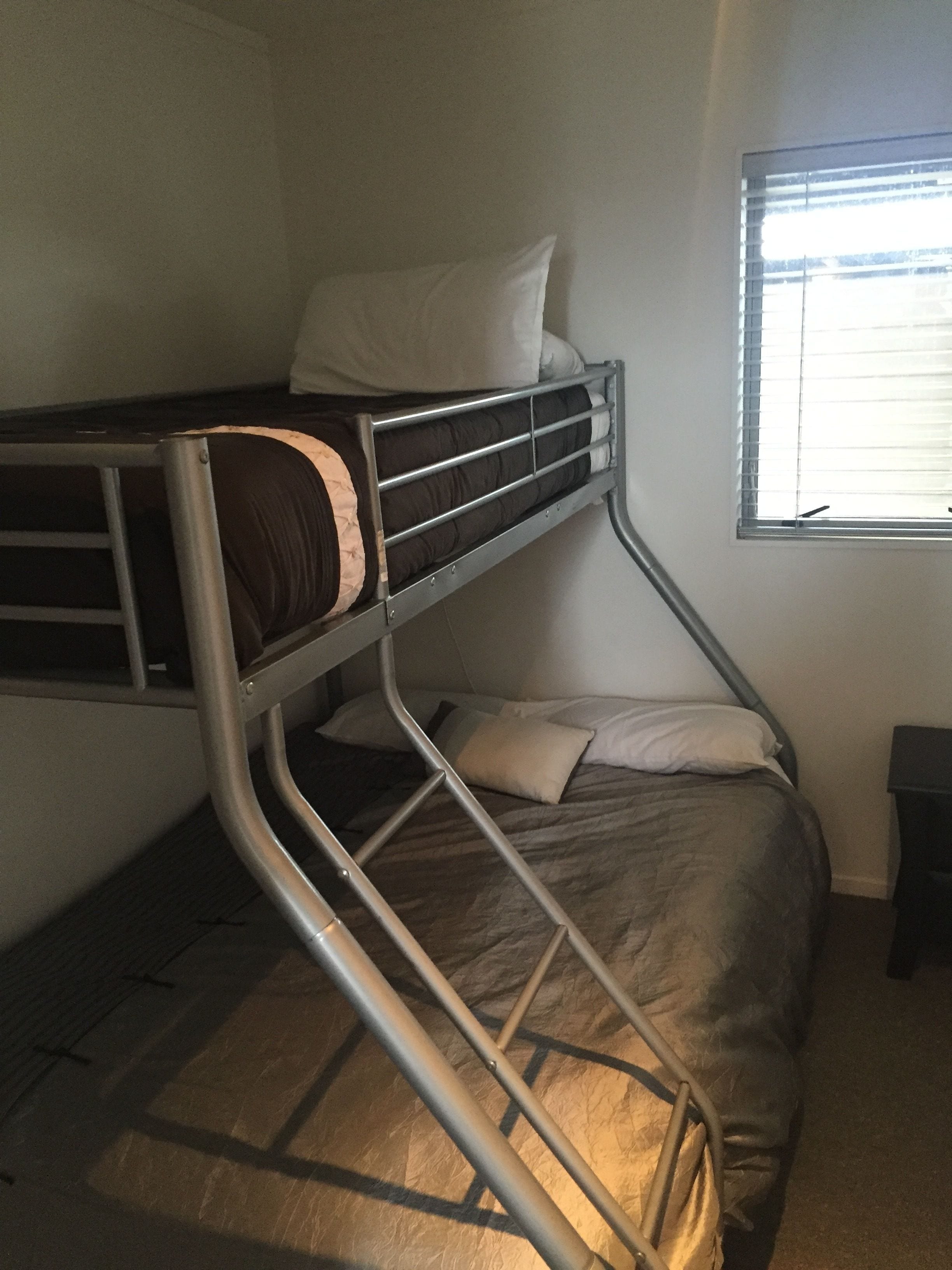 A bunk bed with a metal frame, a single bed below, and a window with blinds.