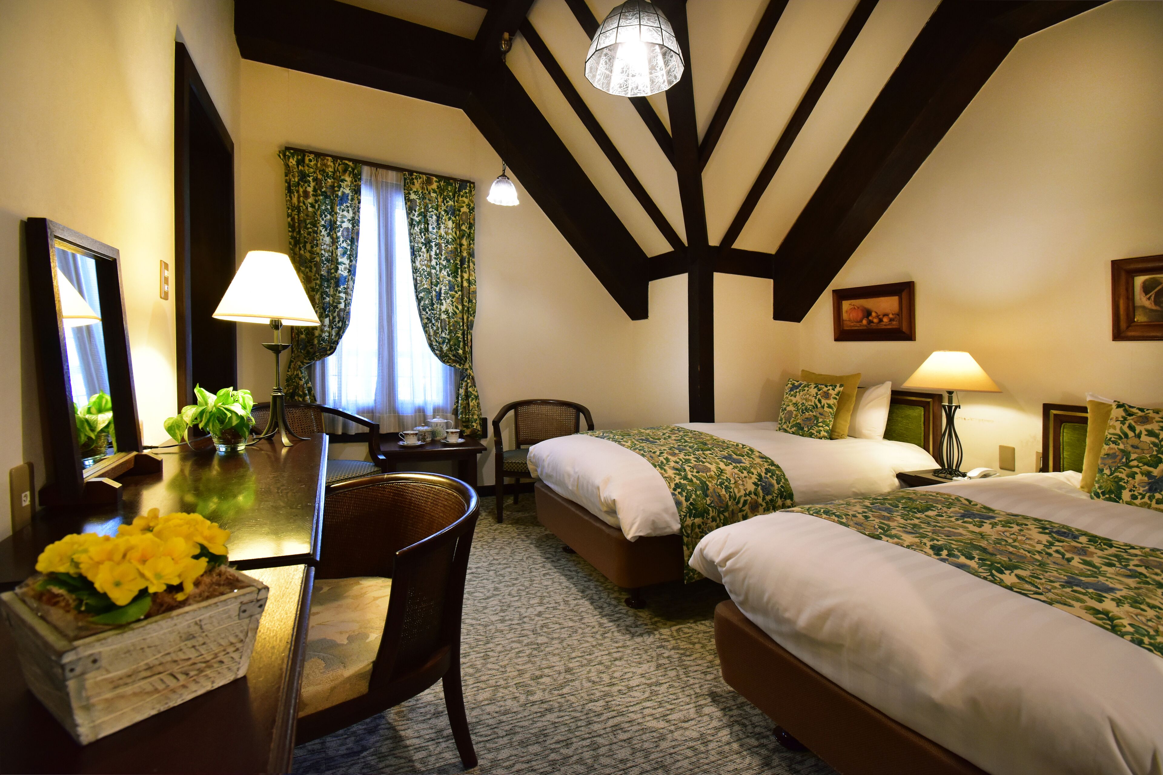A hotel room with two single beds, a desk with a chair, a table with a flower arrangement, and a large window with curtains.