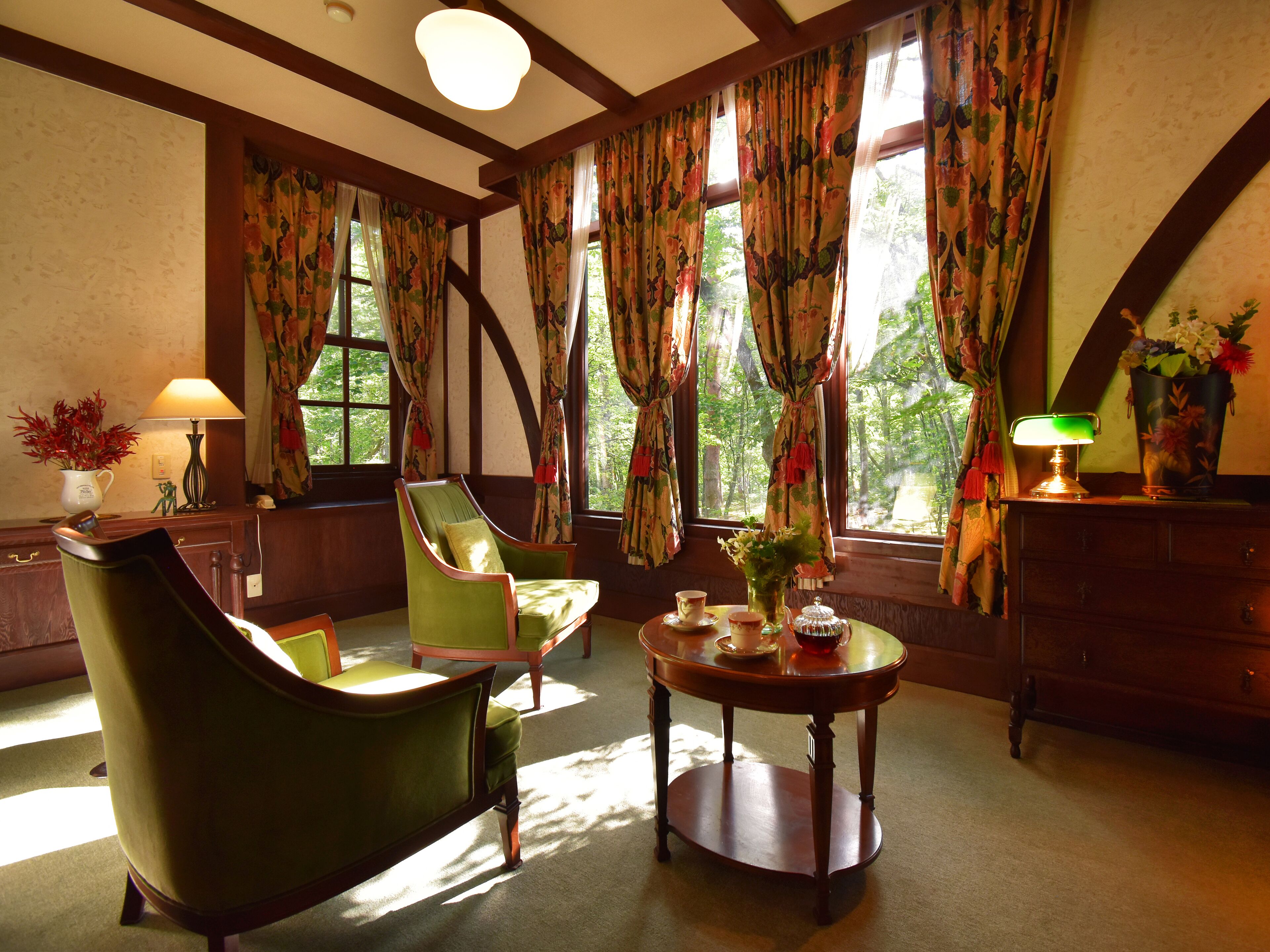 A room with a green armchair, a wooden table with a vase and teapot, and a window with floral curtains.