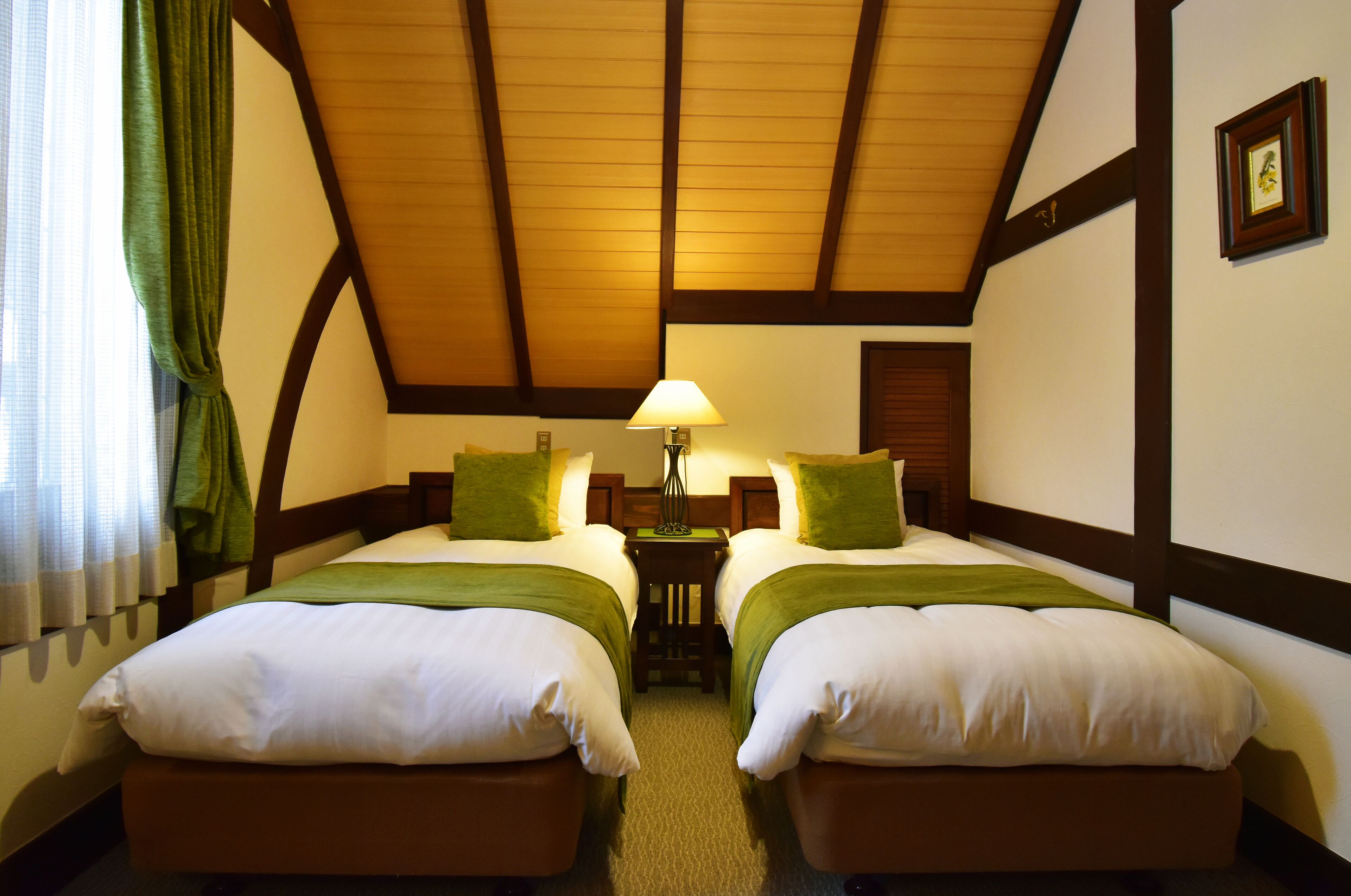 A room with two beds, a wooden ceiling, and a small table with a lamp between them.
