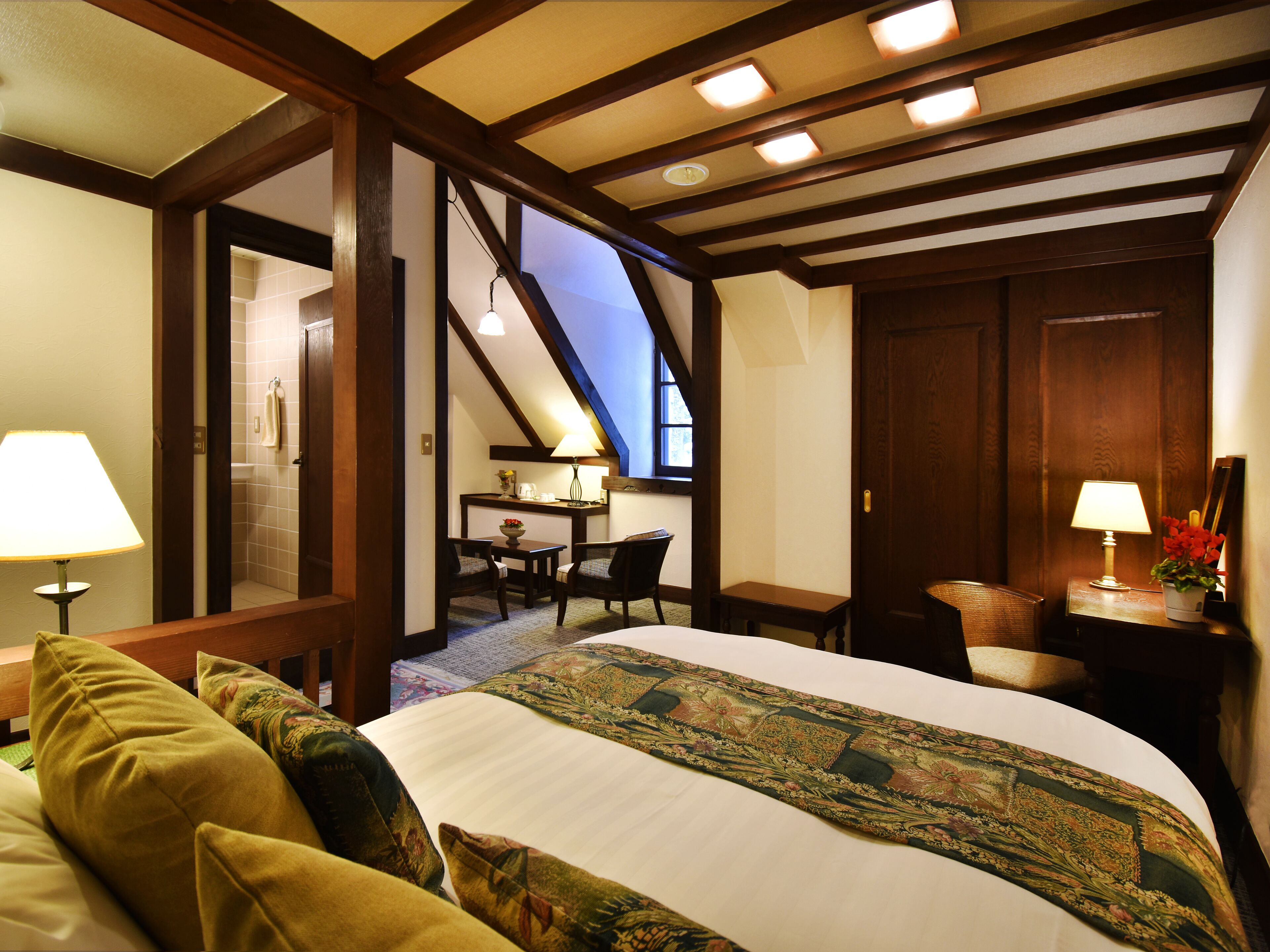 A hotel room with a large bed, a sitting area with a table and chairs, a desk with a lamp, and a view of the sky through a skylight.