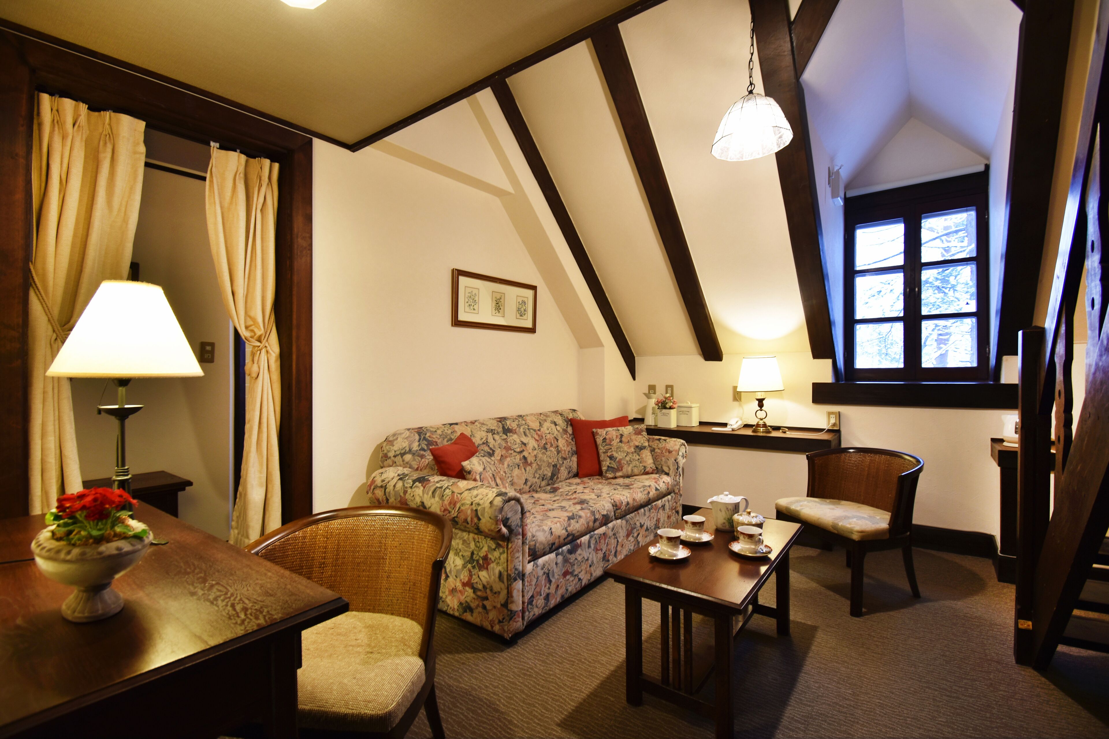 A cozy room with a floral sofa, a dining table with a vase of flowers, and a window with curtains.