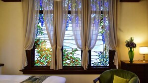 Una habitación con cama, una silla, una mesita de noche con lámpara y un ventanal con vidrieras y cortinas.