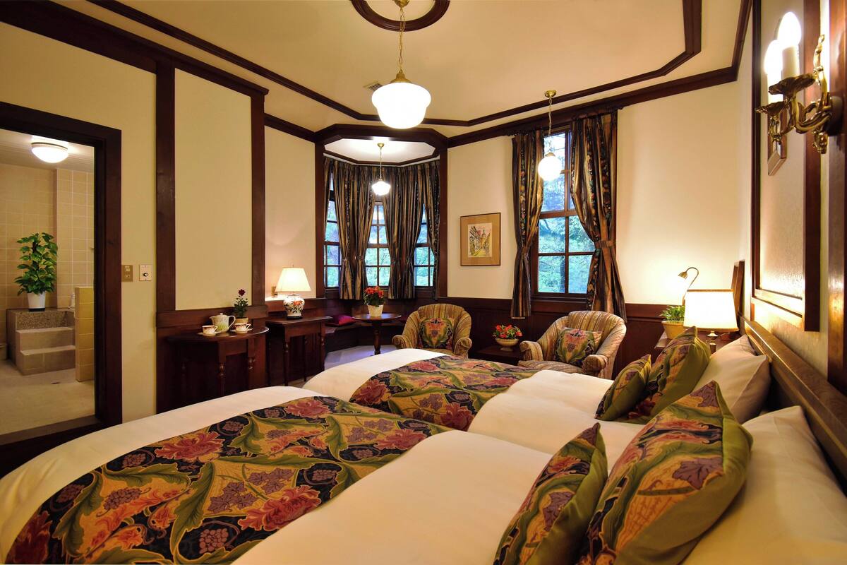 A hotel room with two beds, a desk, a chair, a lamp, and a window with curtains.