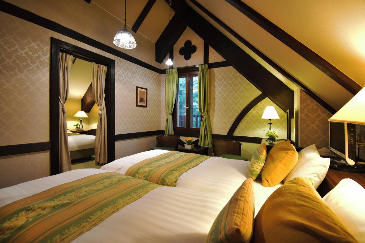 A bedroom with two beds, a window with curtains, a bedside table with a lamp, and a mirror.