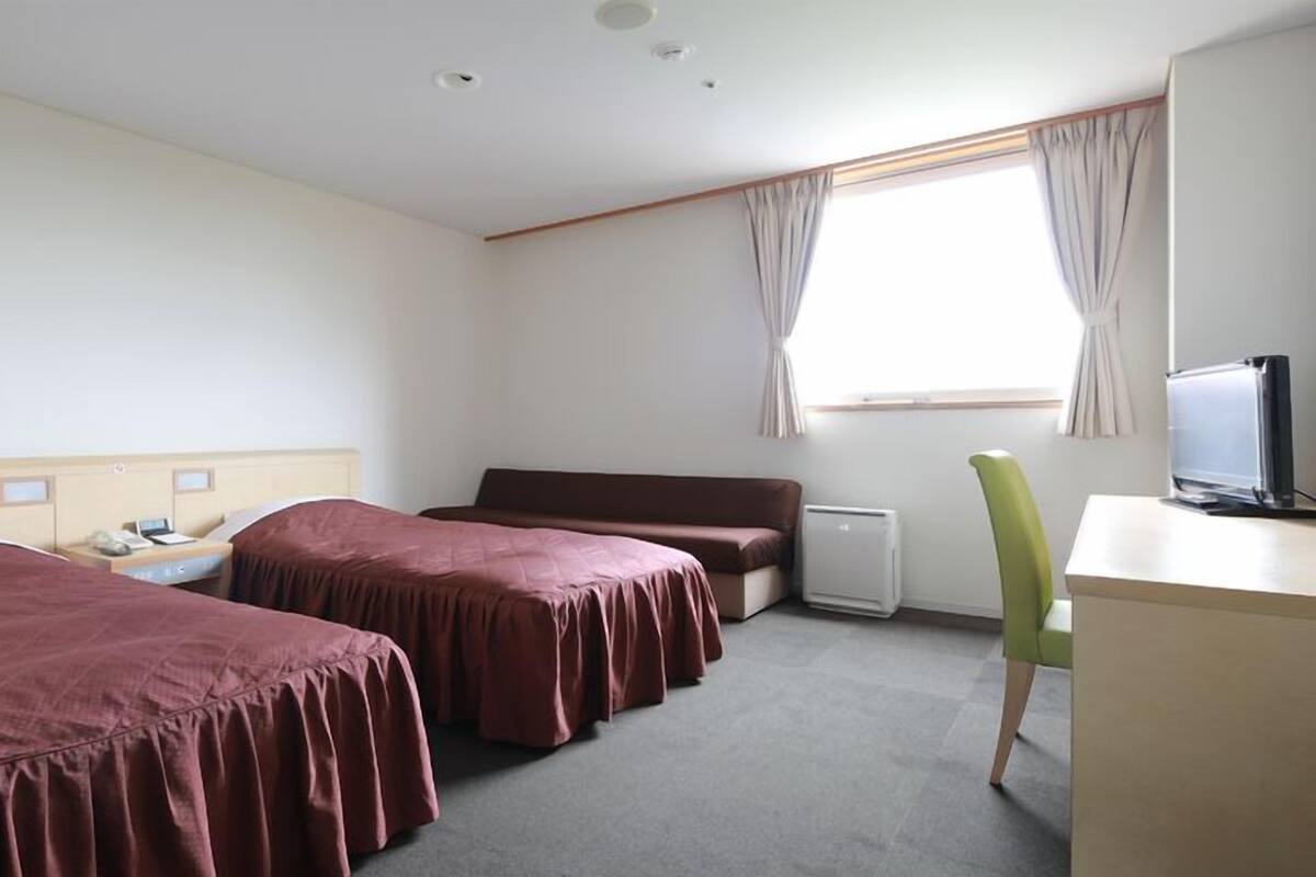 Habitación de hotel con dos camas, un escritorio, una silla, un radiador y una ventana con cortinas.