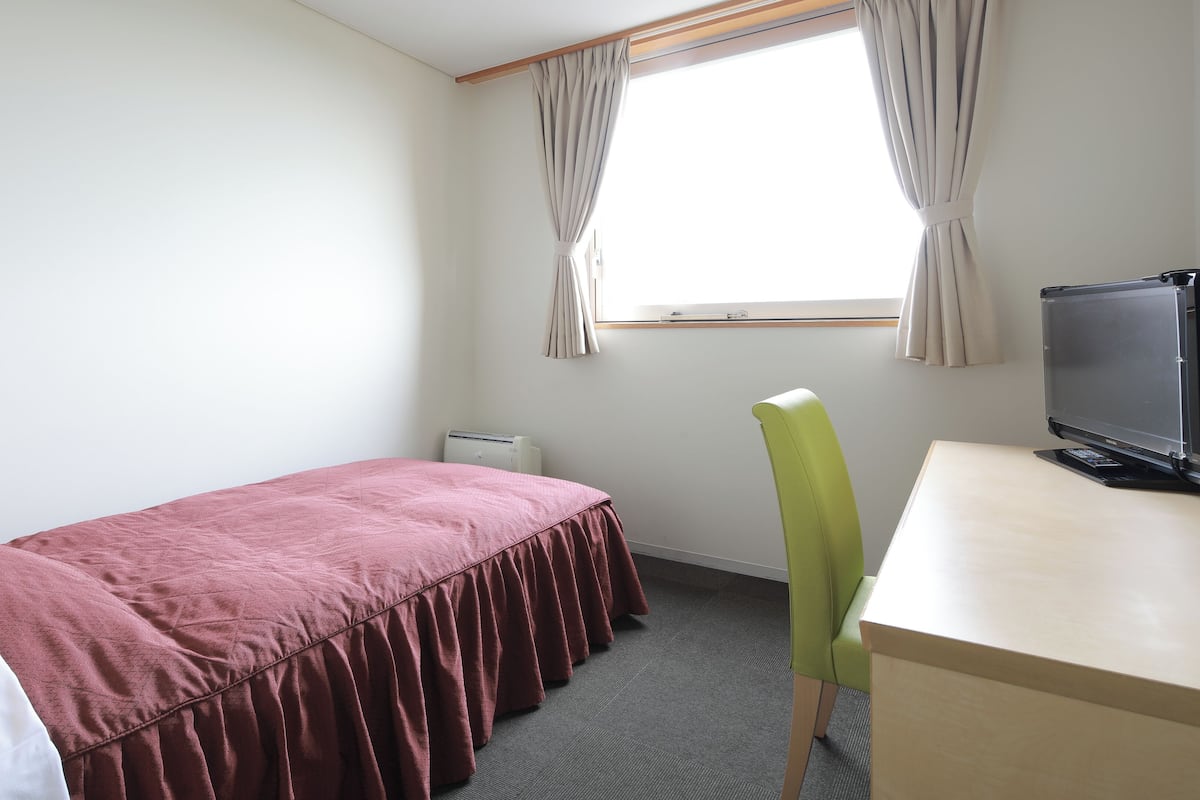 A single bed with a red bedspread, a green chair, a desk with a computer, and a window with curtains.