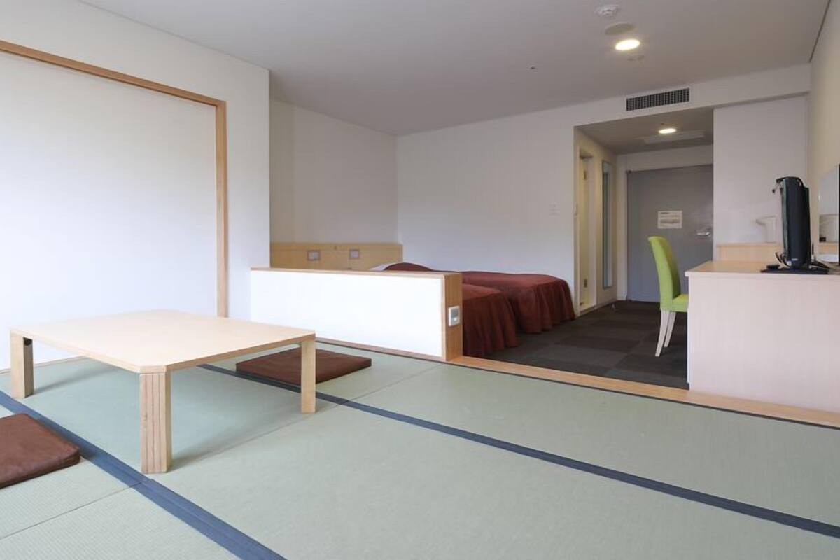 Una habitación con una mesa de madera, un banco, una cama con colcha roja y una silla verde.
