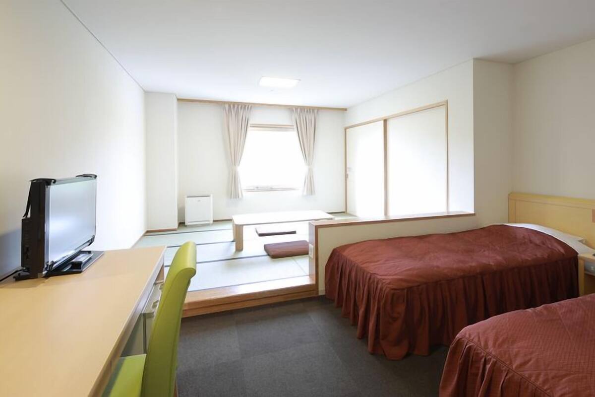Habitación de hotel con dos camas, un escritorio con televisor, una mesita de noche con teléfono y una ventana con cortinas.