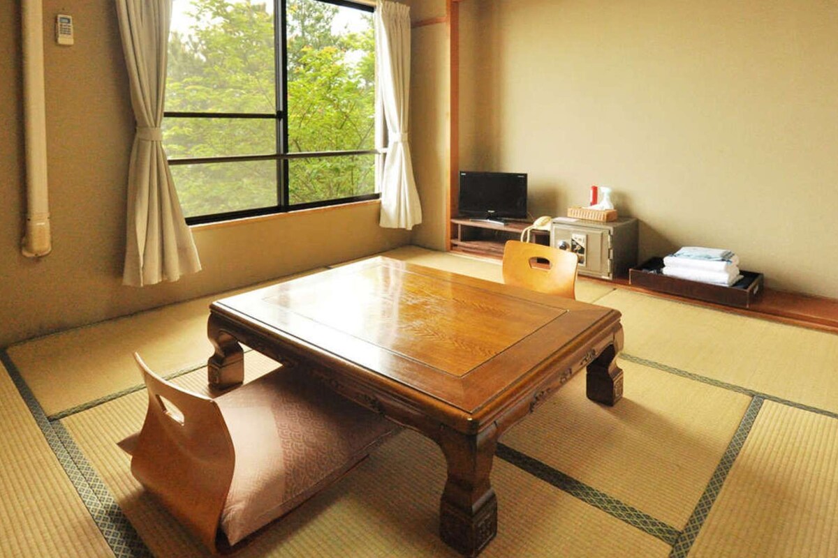 Una habitación tradicional japonesa con suelo de tatami, una mesa de madera baja, un banco acolchado, un televisor y un teléfono.