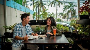 A man and a woman sitting at a table in an outdoor cafe with potted plants and a car in the background. - Hotel 88 Mangga Besar 120 (Jakarta)