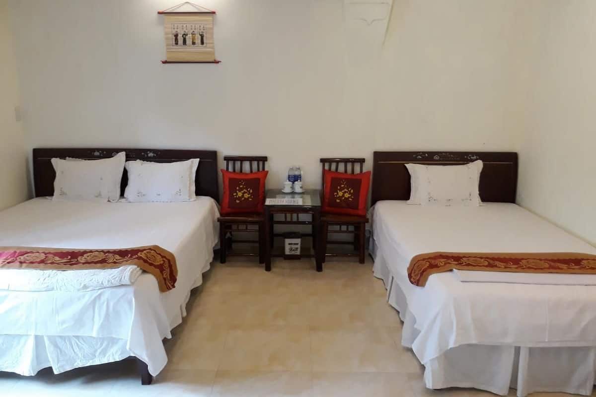 Una habitación con dos camas individuales, cada una con una colcha blanca y un corredor de cama rojo con motivos florales. En el centro hay una pequeña mesa con una botella de agua y una placa de identificación.