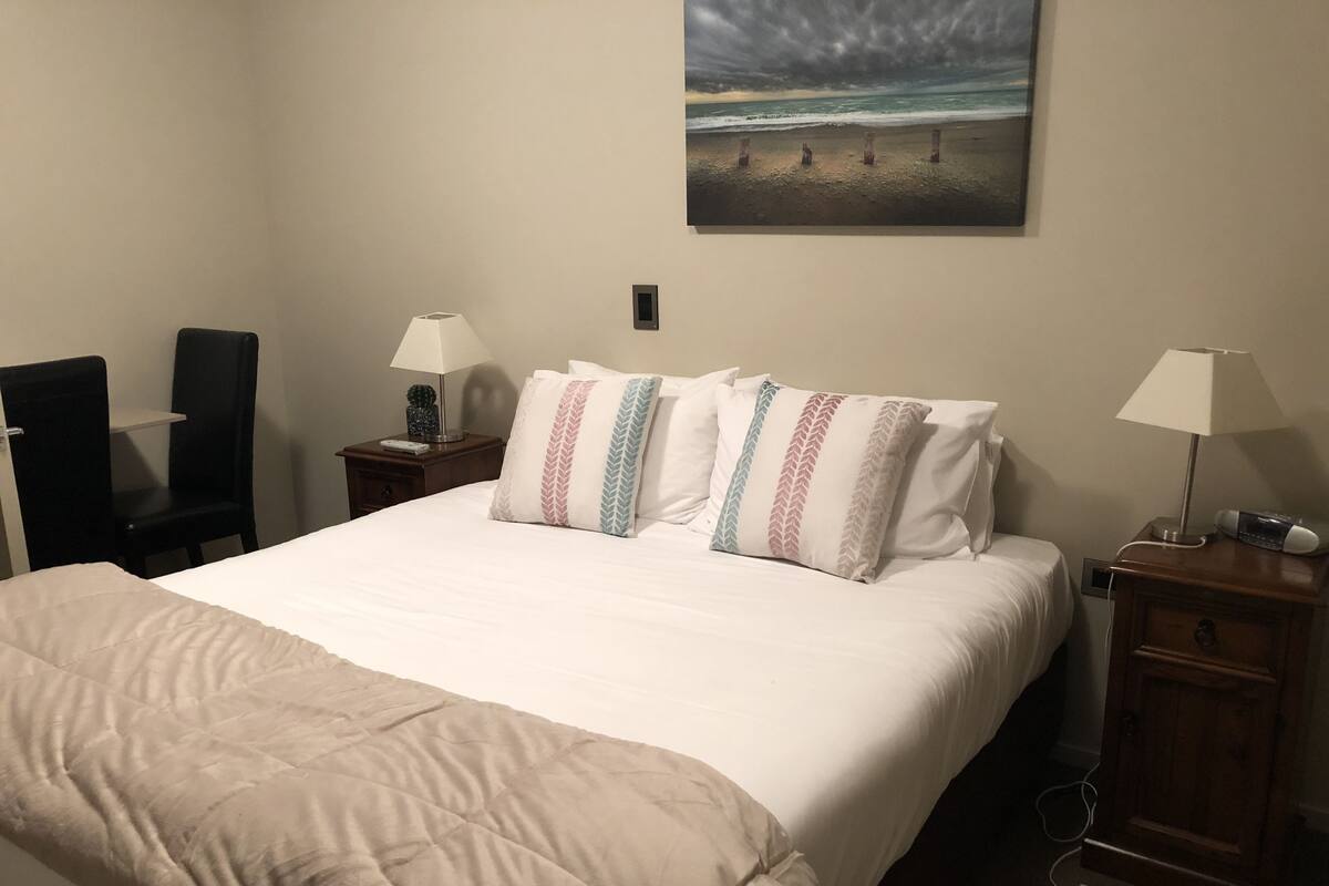 Habitación de hotel con una cama, dos mesitas de noche, una silla, una lámpara y un cuadro de una playa.