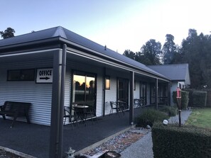 A building with an 'OFFICE' sign, a covered patio with tables and chairs, and a garden area with a bird feeder. - Lake Brunner Longhouse (Moana)