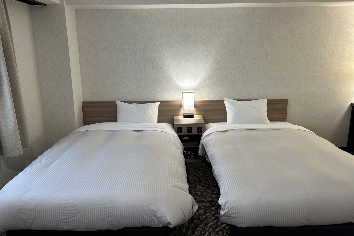 A hotel room with two single beds, a nightstand with a lamp, and a wall-mounted air vent.