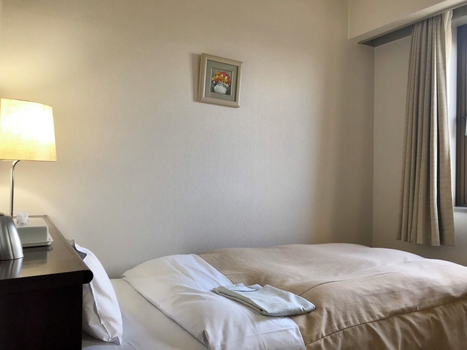 A hotel room with a bed, a bedside table with a lamp, a framed picture on the wall, and a window with curtains.