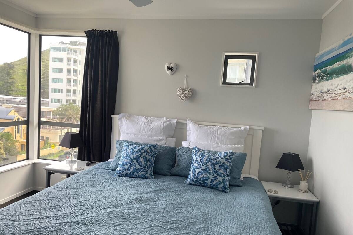 A bedroom with a large bed, a nightstand, a window with curtains, a wall-mounted picture, and a view of buildings outside.