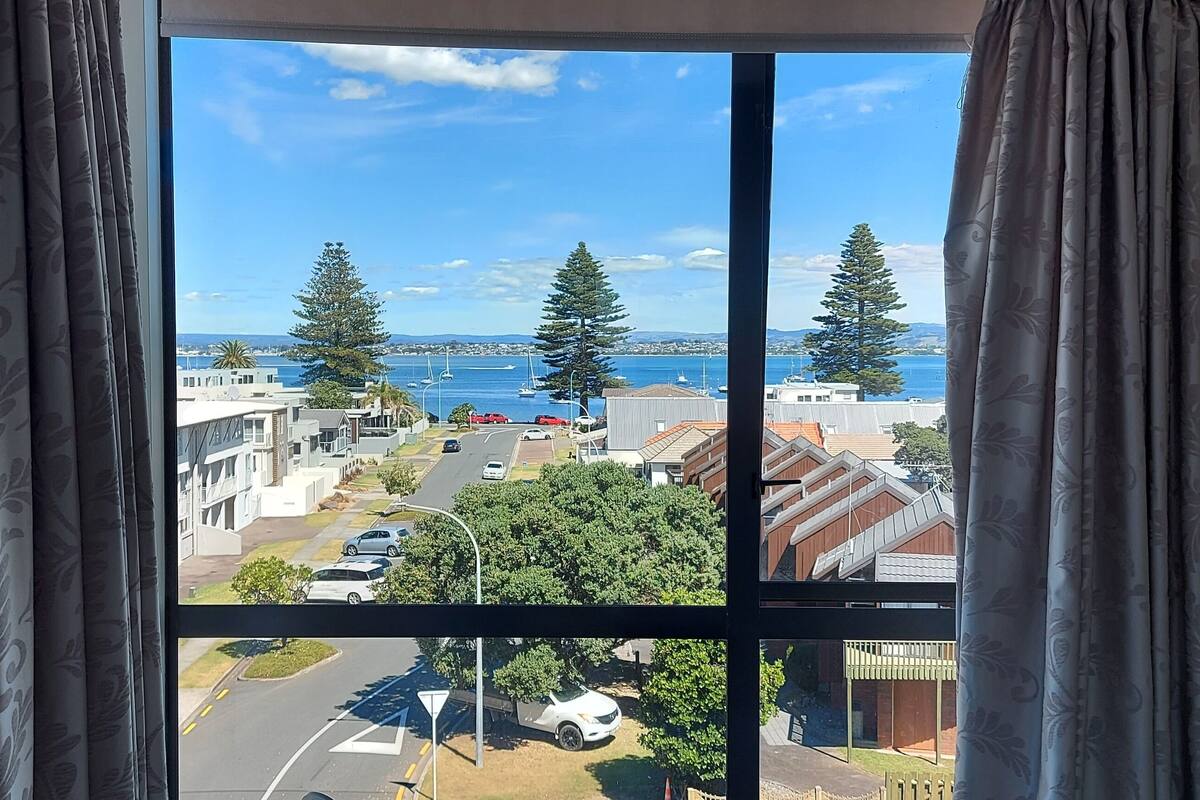 A window with a view of a coastal town, featuring residential buildings, a road, and a body of water.