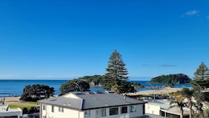 A coastal view with a beach, ocean, and a row of white buildings with a sloped roof. - The Atrium Apartments (Tauranga)