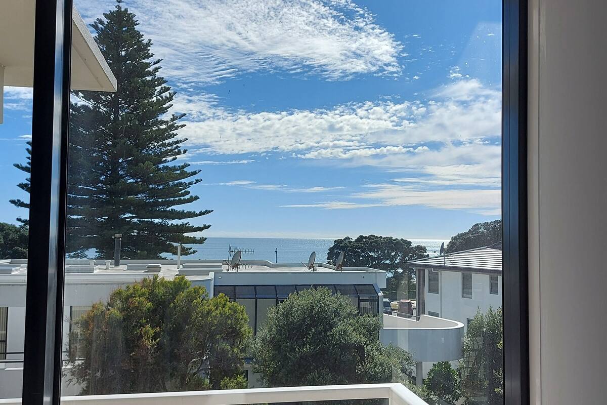A view from a window overlooking a residential area with a clear sky, a large coniferous tree, and a distant body of water.