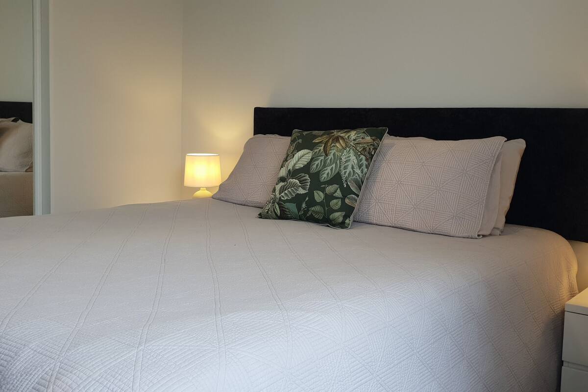 A neatly made bed with a quilted cover, two pillows, a headboard, bedside lamps, and a mirror on the wall.