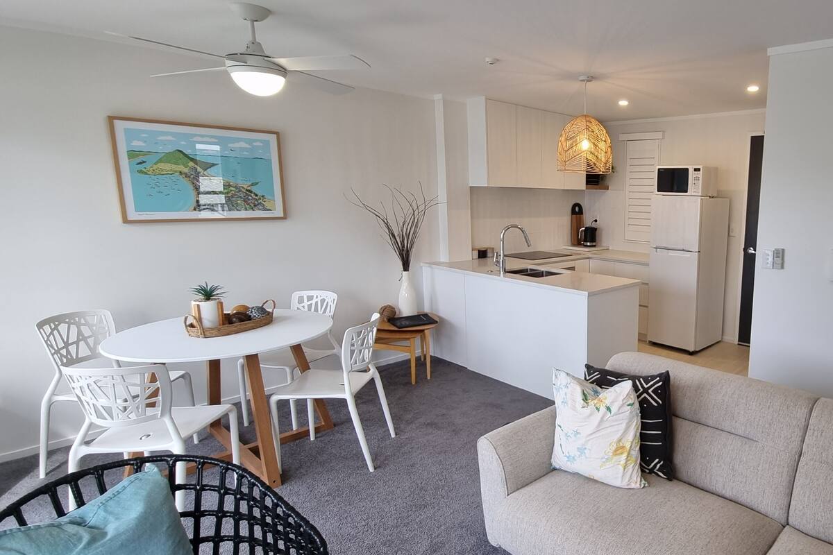 A modern kitchen with white cabinets, a microwave, and a refrigerator. There is a dining area with a round table and white chairs. A sofa with patterned cushions is in the foreground.