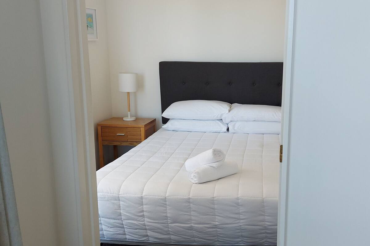 A neatly made bed with white bedding and pillows, a bedside lamp, and a wooden nightstand.