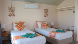 A hotel room with two beds, each with orange and white bedding, a small bedside table with a lamp, and decorative wall hangings.